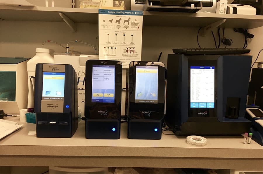 Three veterinary diagnostic machines with touchscreens are displayed on a countertop in a lab, with various supplies and a sample handling guide visible in the background.