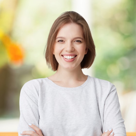 A person with short brown hair wearing a light gray sweater smiles with arms crossed, standing in front of a blurred background with green and yellow tones.